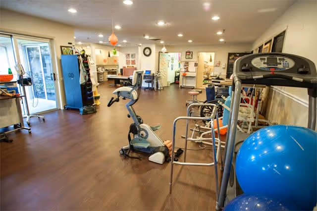 A spacious indoor rehabilitation room with exercise bikes, a treadmill, walkers, and large blue exercise balls.