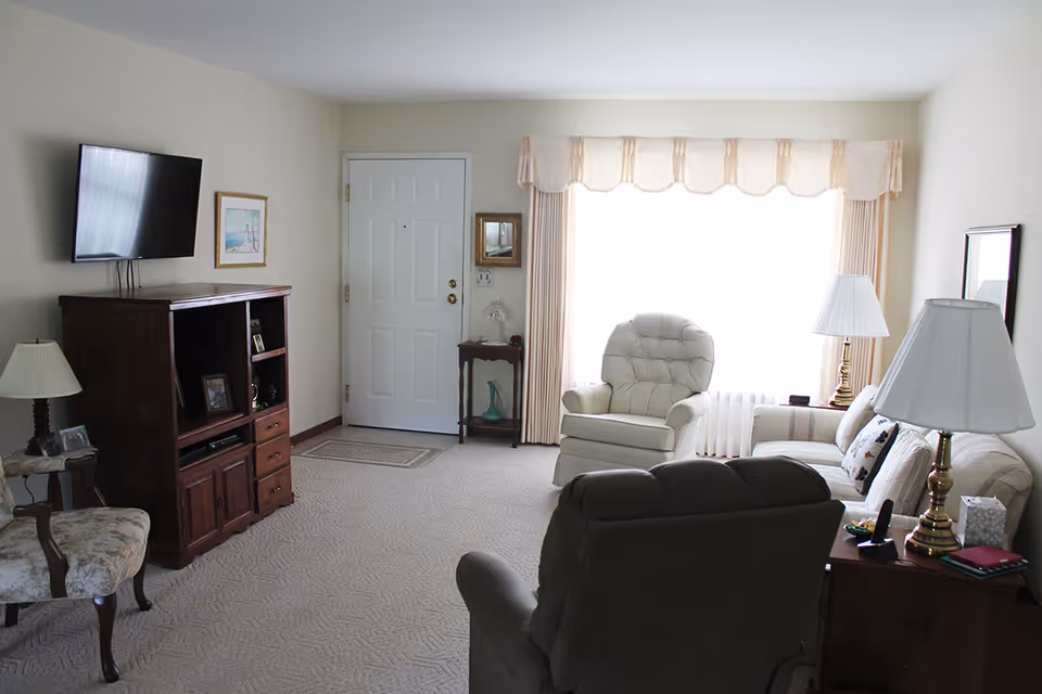 A cozy living room in Good Samaritan Home featuring a large window with beige curtains, a white cushioned armchair, a beige sofa with pillows, two table lamps with white shades, a wooden entertainment center with a mounted flat-screen TV, and additional seating including a floral upholstered chair and a recliner.