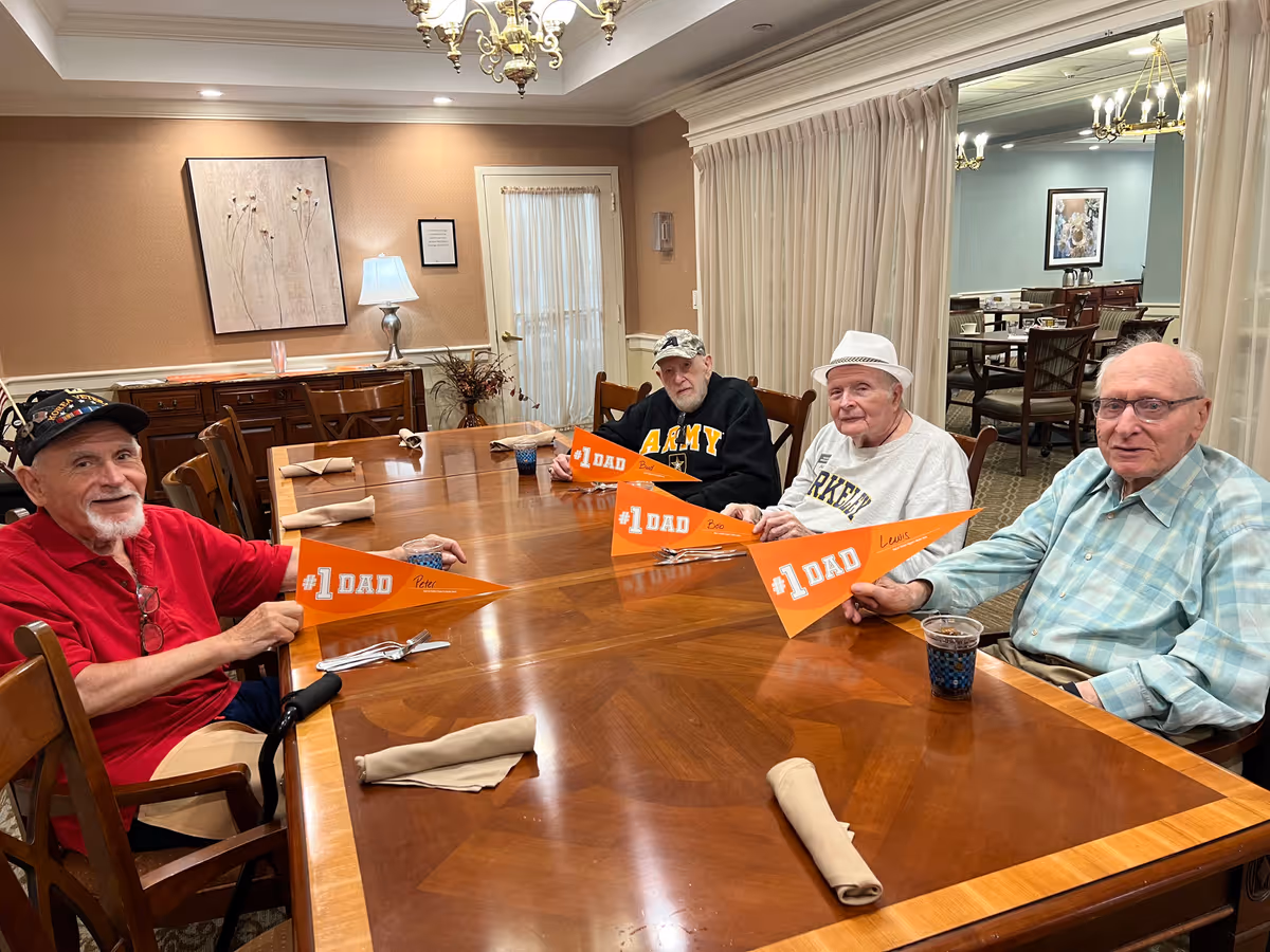Four elderly men sitting around a dining table in a communal dining room holding orange "#1 DAD" pennants.