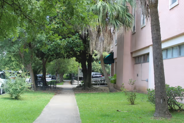 Sidewalk running alongside a pink brick building with green grass, small bushes, and several trees including palm trees. There are parked cars visible on the left side and a bench under the trees.
