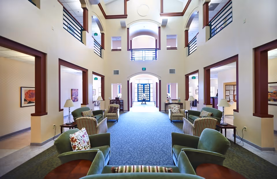 A spacious and well-lit common area in a senior living facility with high ceilings and large windows. The room features multiple seating arrangements with green and striped upholstered chairs and sofas, small wooden tables with lamps, and framed artwork on the walls. The area has an open layout with archways and a carpeted floor, leading to other rooms and a bright entrance in the background.