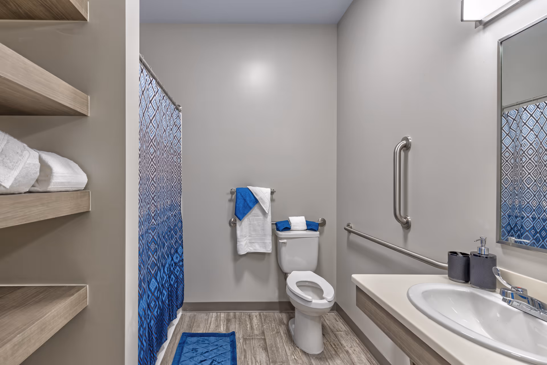 A clean and modern bathroom with a toilet, sink, and shower. The shower has a blue and white patterned curtain. There are blue and white towels hanging on a rack above the toilet and a blue bath mat on the floor. The walls are painted light gray, and there are wooden shelves with folded white towels on the left side. A grab bar is installed next to the toilet and sink for accessibility.