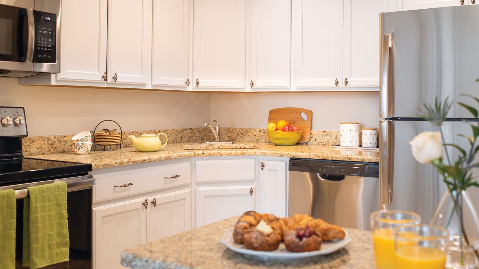 A modern kitchen with white cabinets, granite countertops, and stainless steel appliances including a refrigerator, dishwasher, microwave, and stove. There are green towels hanging on the oven handle, a yellow teapot and mugs on the counter, a bowl of fruit, and a plate of pastries on the kitchen island. A vase with a white flower is also visible on the island.