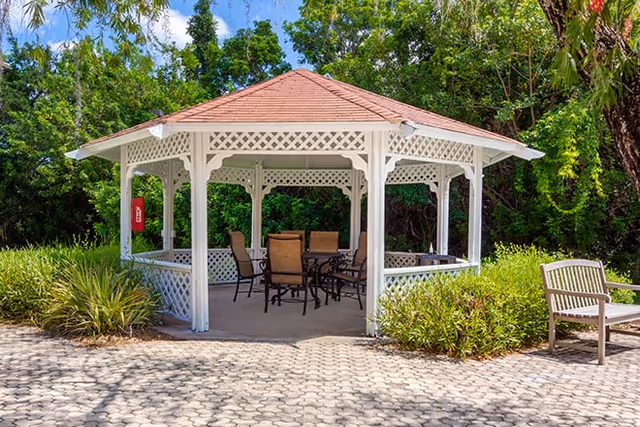 White octagonal gazebo with a patio table and chairs surrounded by landscaping and a bench.