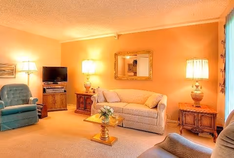 A cozy living room with beige walls and carpet, featuring a beige sofa with cushions, two wooden side tables each with a lamp, a wooden coffee table with a flower arrangement, a green armchair, a TV on a wooden stand, and a large mirror hanging on the wall.