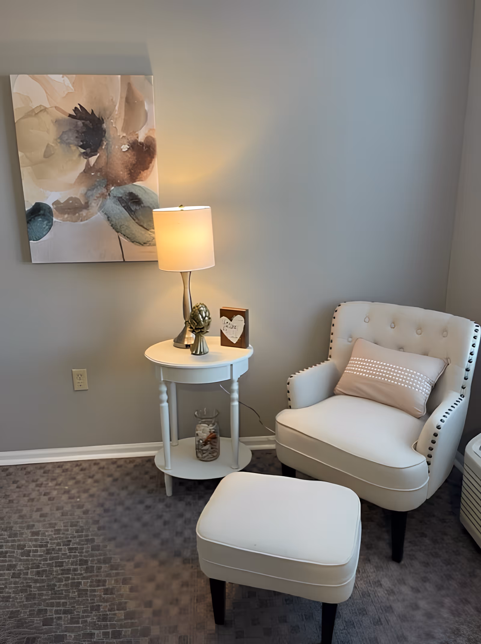 A cozy corner with a white upholstered armchair and matching ottoman on a patterned carpet. Next to the chair is a small round white side table with a lamp, decorative artichoke, and a small framed sign. A large abstract floral painting hangs on the light gray wall behind the table.