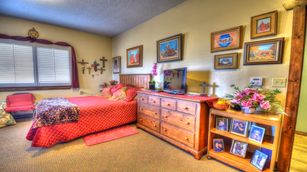 A cozy bedroom with a red quilted bed, wooden dresser topped with flowers and a TV, and framed pictures and crosses on the walls.