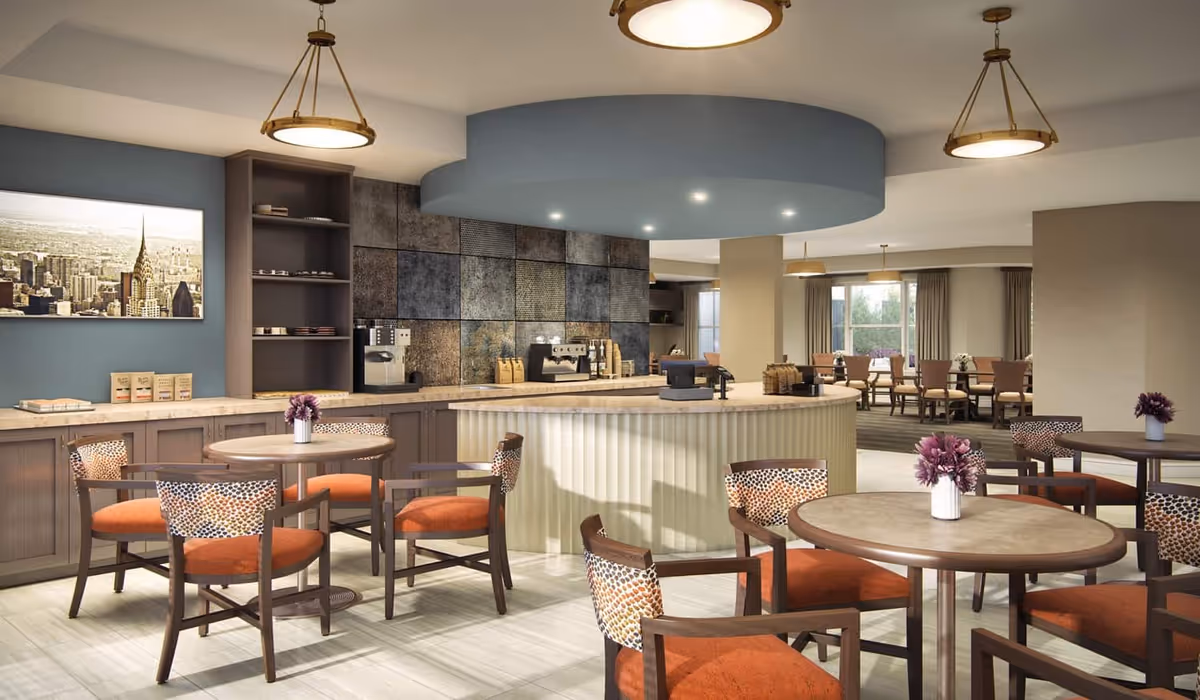 Bright communal dining and café area with round tables, orange-upholstered chairs and a service counter with coffee machines.