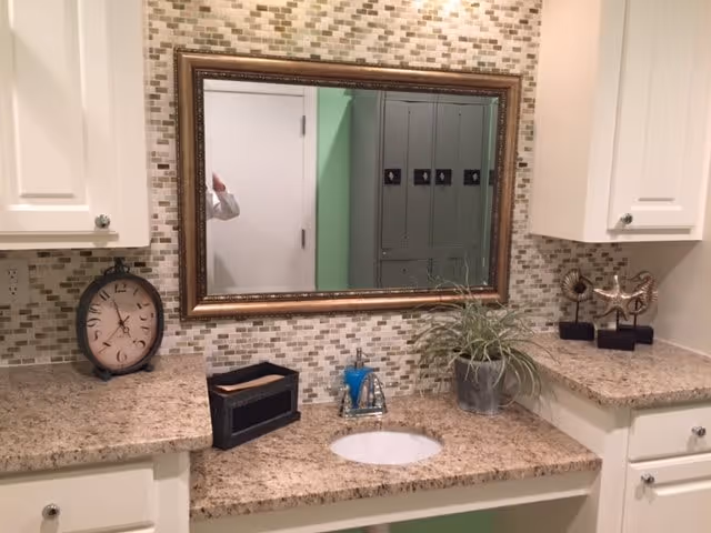 A bathroom vanity with a granite countertop, an oval sink, a silver faucet, and a large rectangular mirror with a decorative frame. The backsplash is made of small mosaic tiles in neutral tones. On the countertop, there is a round clock, a black tissue box holder, a potted plant, and two decorative starfish sculptures. White cabinets are mounted on the wall above the countertop.