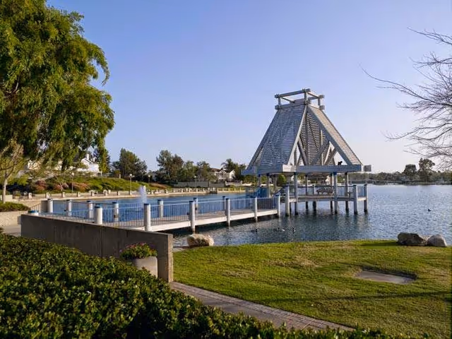 A wooden pavilion on a dock extending over a calm lake with grassy shore, trees, and nearby homes under a clear sky.