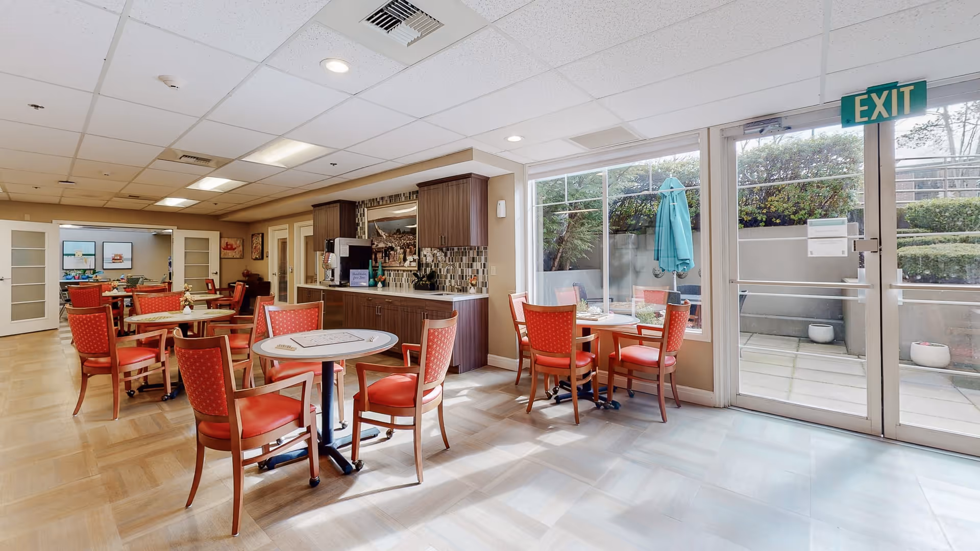A bright dining area in Island House Assisted Living with several round tables and red cushioned chairs. There is a small kitchenette with cabinets, a coffee machine, and a tiled backsplash. Large windows and a glass door provide natural light and a view of an outdoor patio with greenery and an umbrella-covered table. The floor is a mix of light wood and tile, and there is an exit sign above the door.