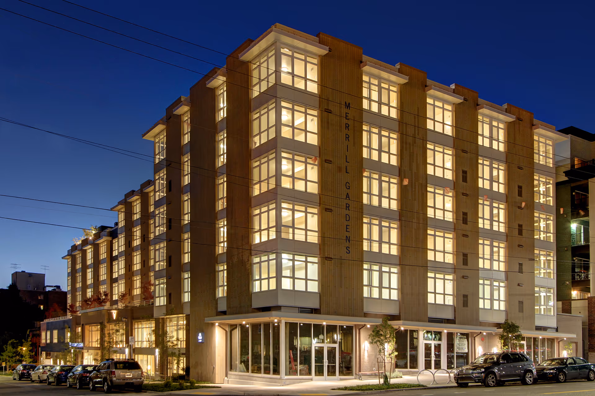 Exterior nighttime view of a multi-story building with large illuminated windows. The building has the name Merrill Gardens displayed vertically on the corner. Several cars are parked along the street in front of the building.