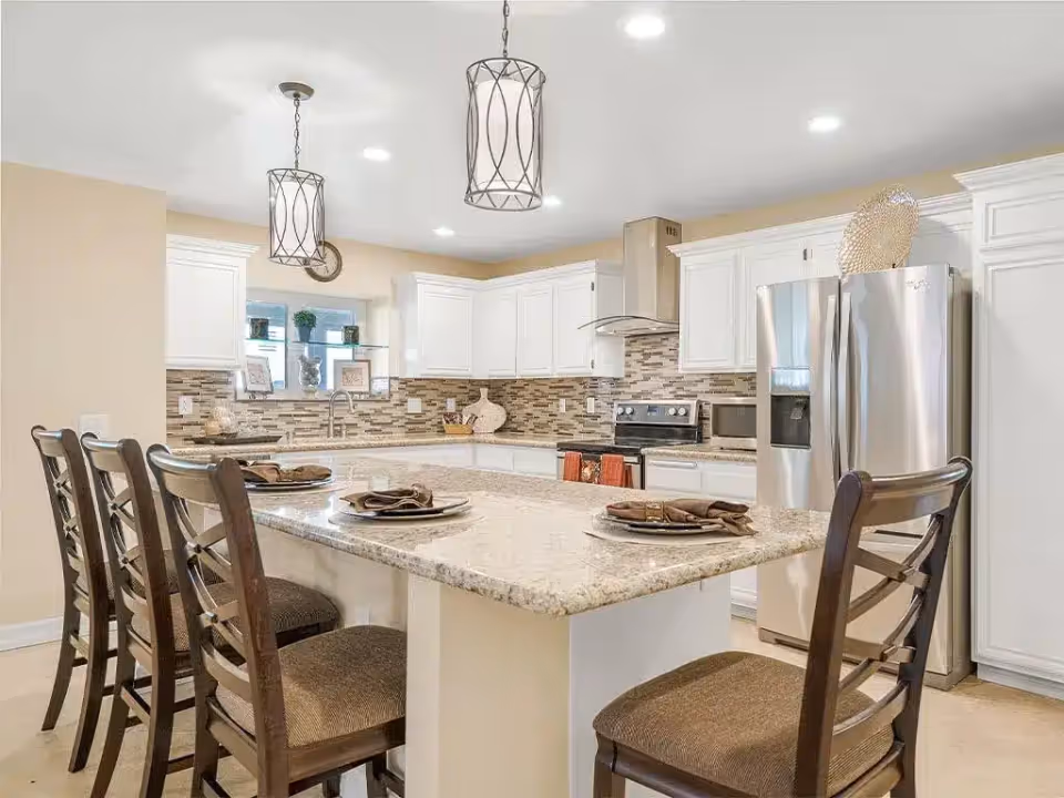 A modern kitchen with white cabinets, a stainless steel refrigerator, oven, and microwave. There is a large granite island countertop with four wooden chairs, each set with a plate and napkin. Two decorative pendant lights hang from the ceiling, and a tiled backsplash runs along the walls behind the counters.