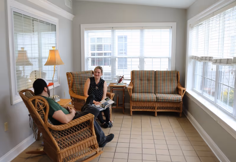 A bright room with large windows and tiled floor featuring wicker furniture including a rocking chair, armchair, and loveseat with plaid cushions. A man and woman are seated and engaged in conversation, with the woman holding a magazine. A table with a lamp and decorative items is placed between the chairs.