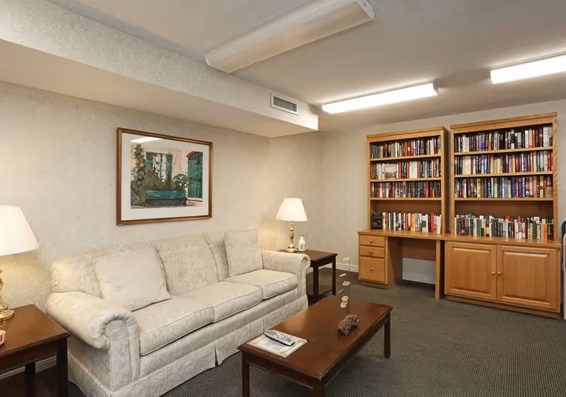 A cozy living room with a beige patterned sofa, two wooden side tables each with a lamp, a wooden coffee table with a remote and a small decorative item, and two wooden bookshelves filled with books against the wall. A framed picture hangs above the sofa.