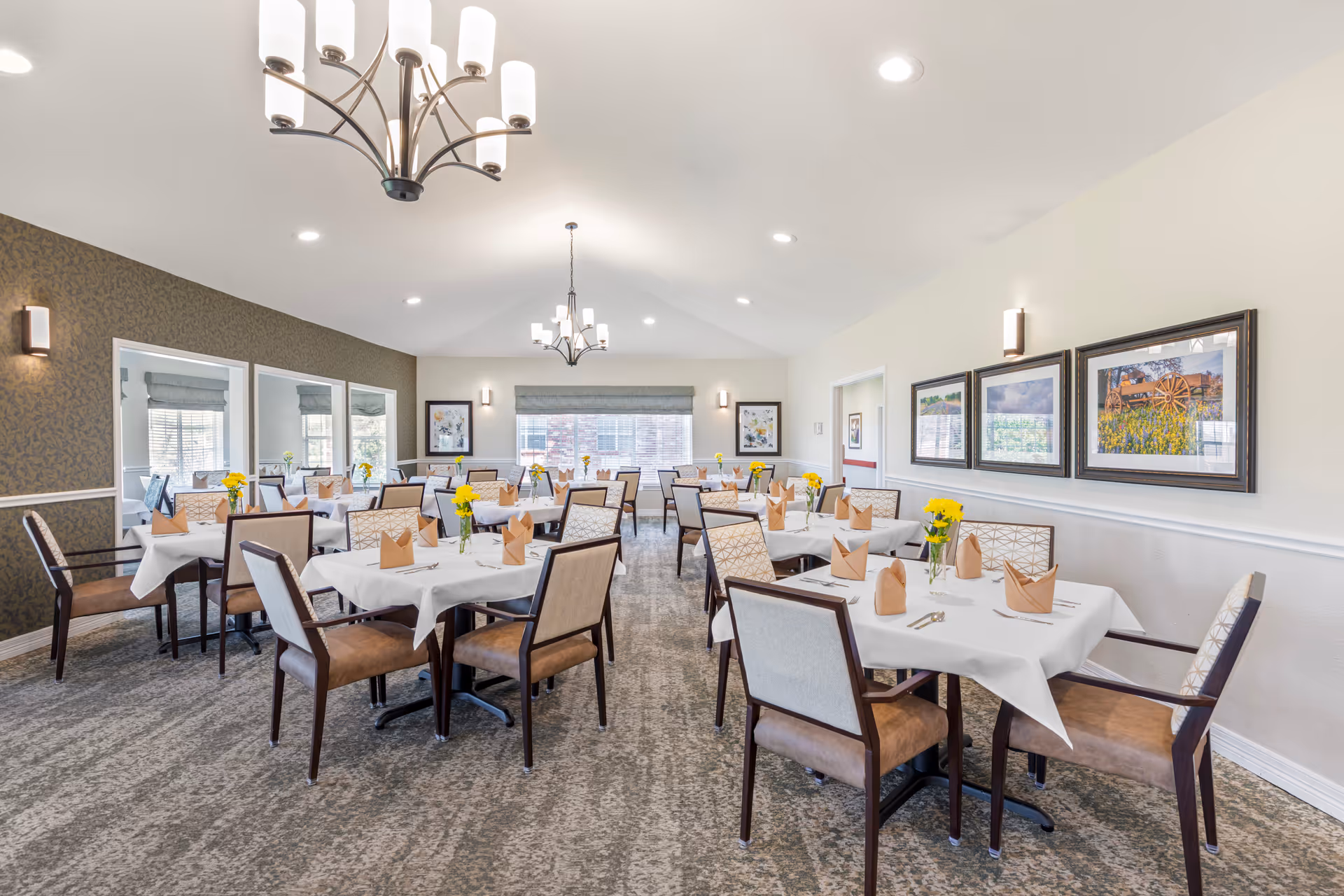A bright dining room with multiple tables set with white tablecloths, folded napkins and yellow flowers at a senior living facility.