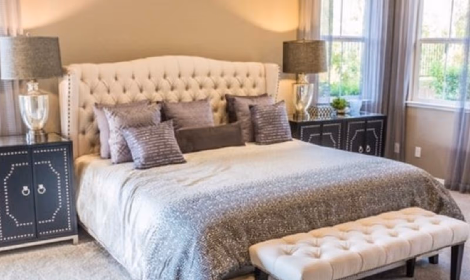 A well-decorated bedroom featuring a large tufted headboard bed with multiple decorative pillows, two matching black nightstands with silver lamps, sheer curtains on windows letting in natural light, and a tufted bench at the foot of the bed.