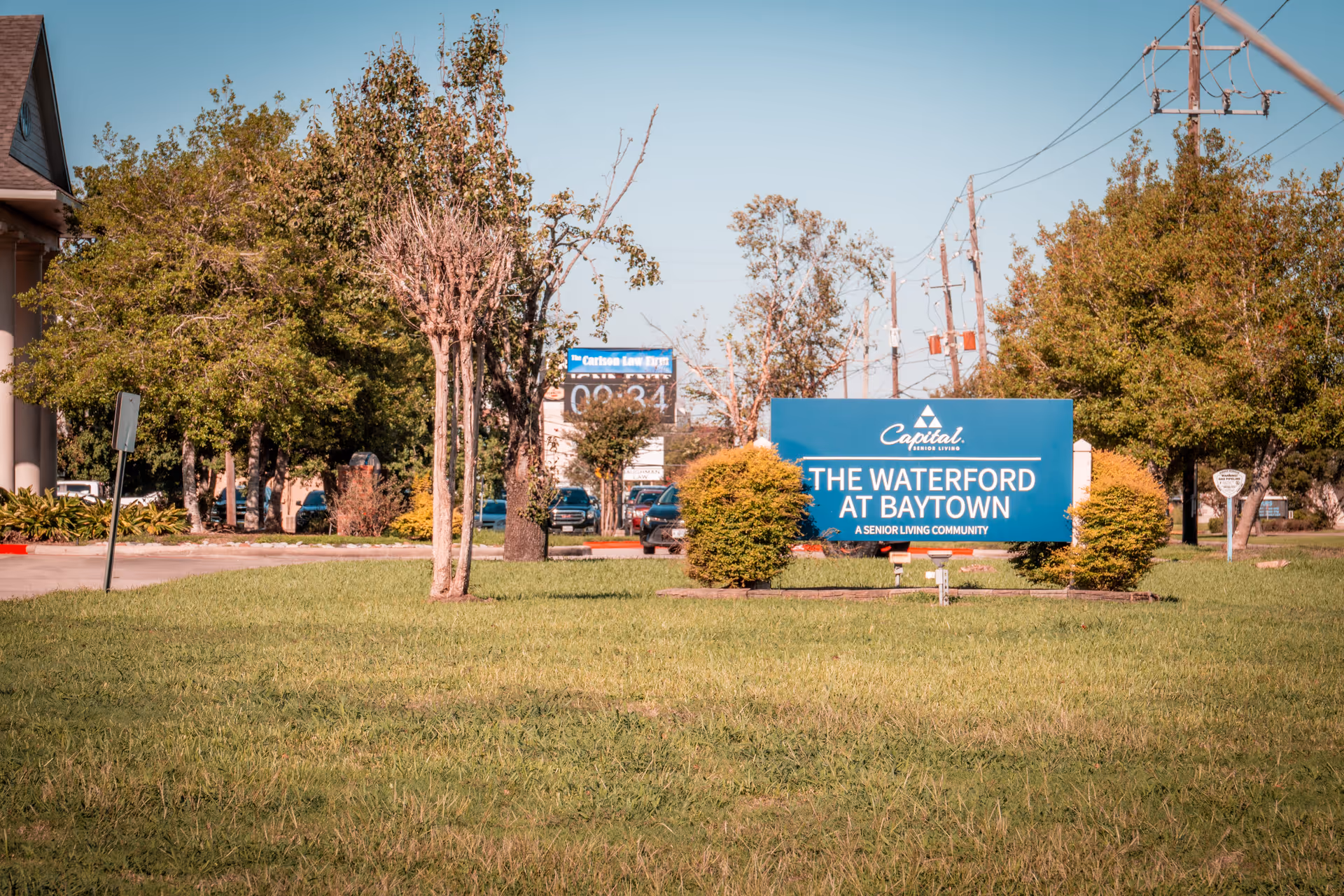 Entrance sign reading "The Waterford at Baytown" on a grassy lawn with trees, nearby cars, and utility poles in the background.