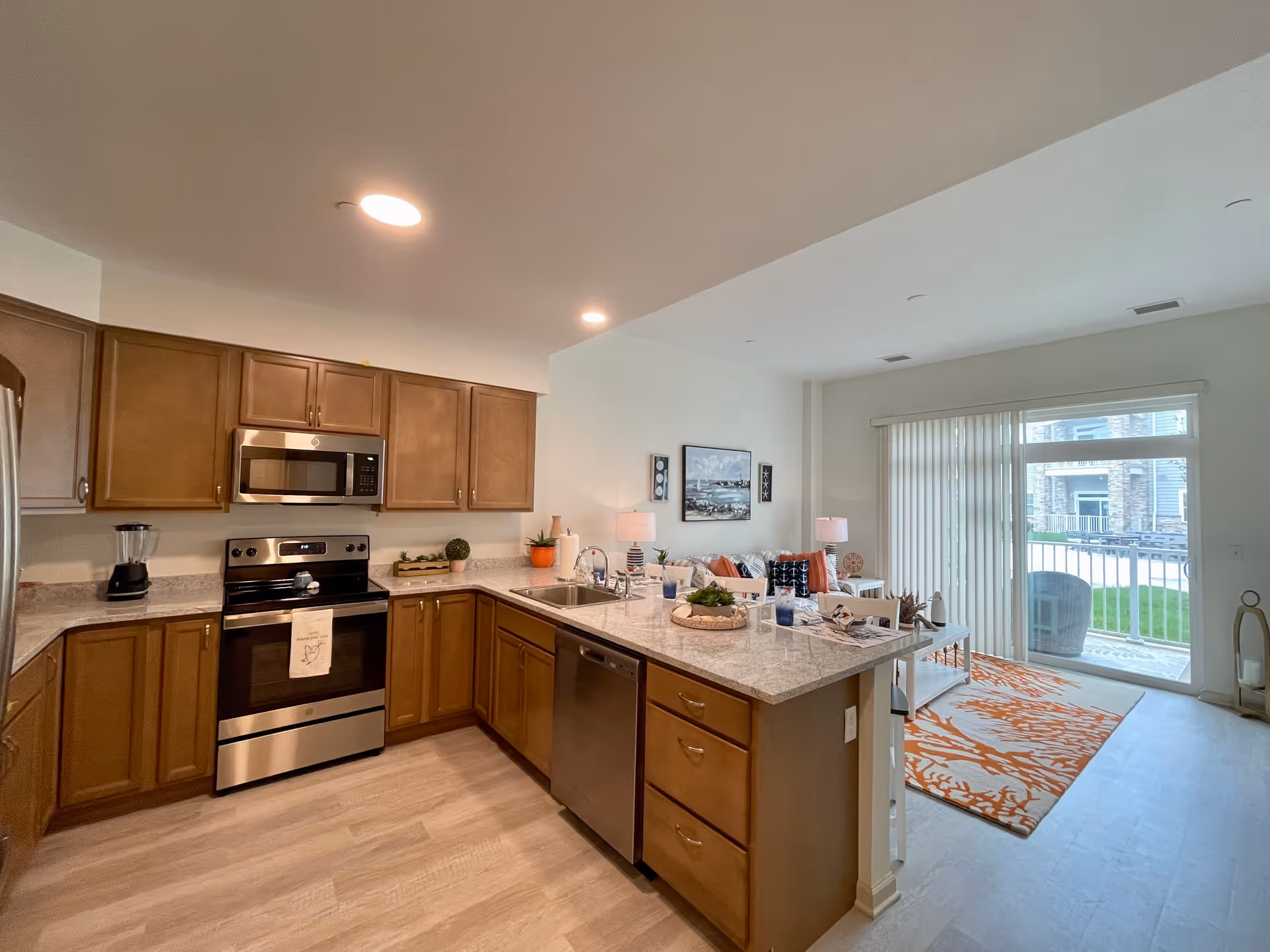 Open-plan kitchen with wooden cabinets, stainless steel appliances and a marble-topped island opening to a living area with a sliding glass door.
