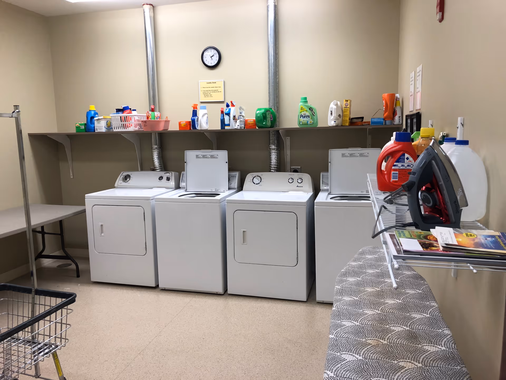 Laundry room with two washing machines and two dryers lined up against a beige wall. Above the machines is a shelf holding various laundry detergents and cleaning supplies. A clock and a small yellow sign are mounted on the wall above the shelf. To the right, there is an ironing board with an iron and some magazines on a wire rack. A laundry cart is partially visible in the foreground on the left side.