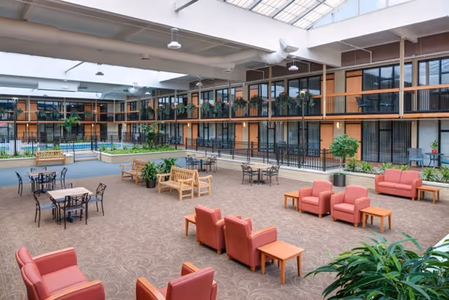 Spacious indoor common area with seating arrangements including red armchairs, wooden benches, and tables with chairs. The area is carpeted and surrounded by two-story walls with large windows and plants. A glass ceiling allows natural light to illuminate the space.