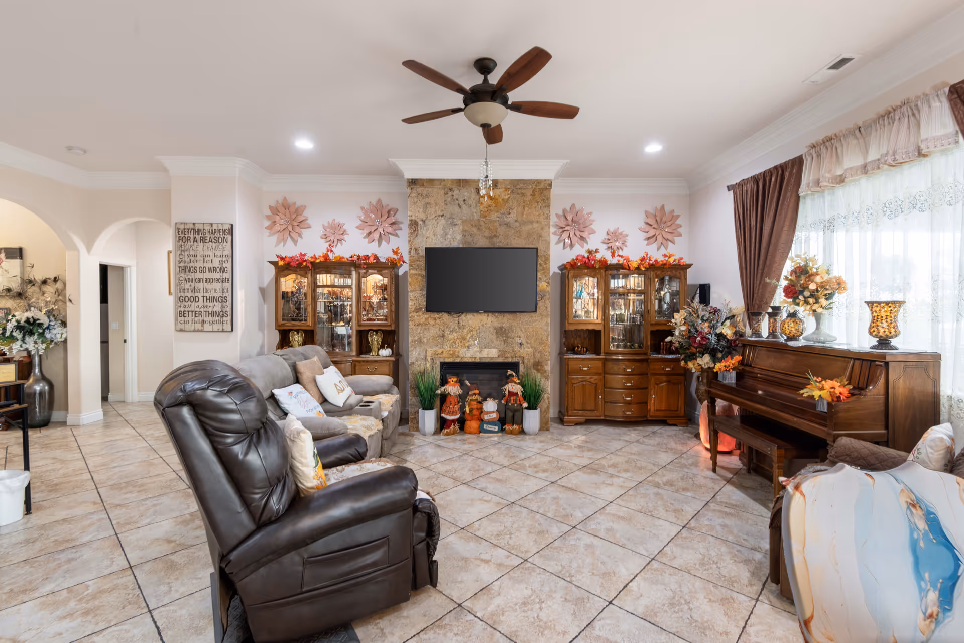 Bright, decorated living room with a recliner and sofa facing a wall-mounted TV above a stone fireplace, flanked by display cabinets and a piano by the window.