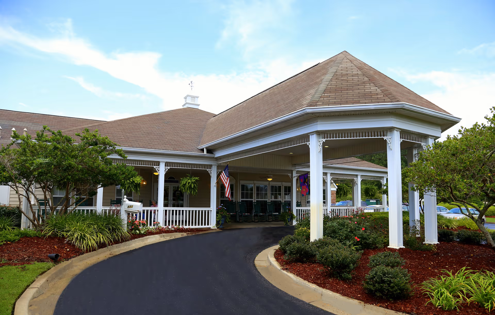 Exterior view of a senior living facility entrance with a covered driveway, white pillars, landscaped bushes and trees, an American flag, and a clear blue sky.