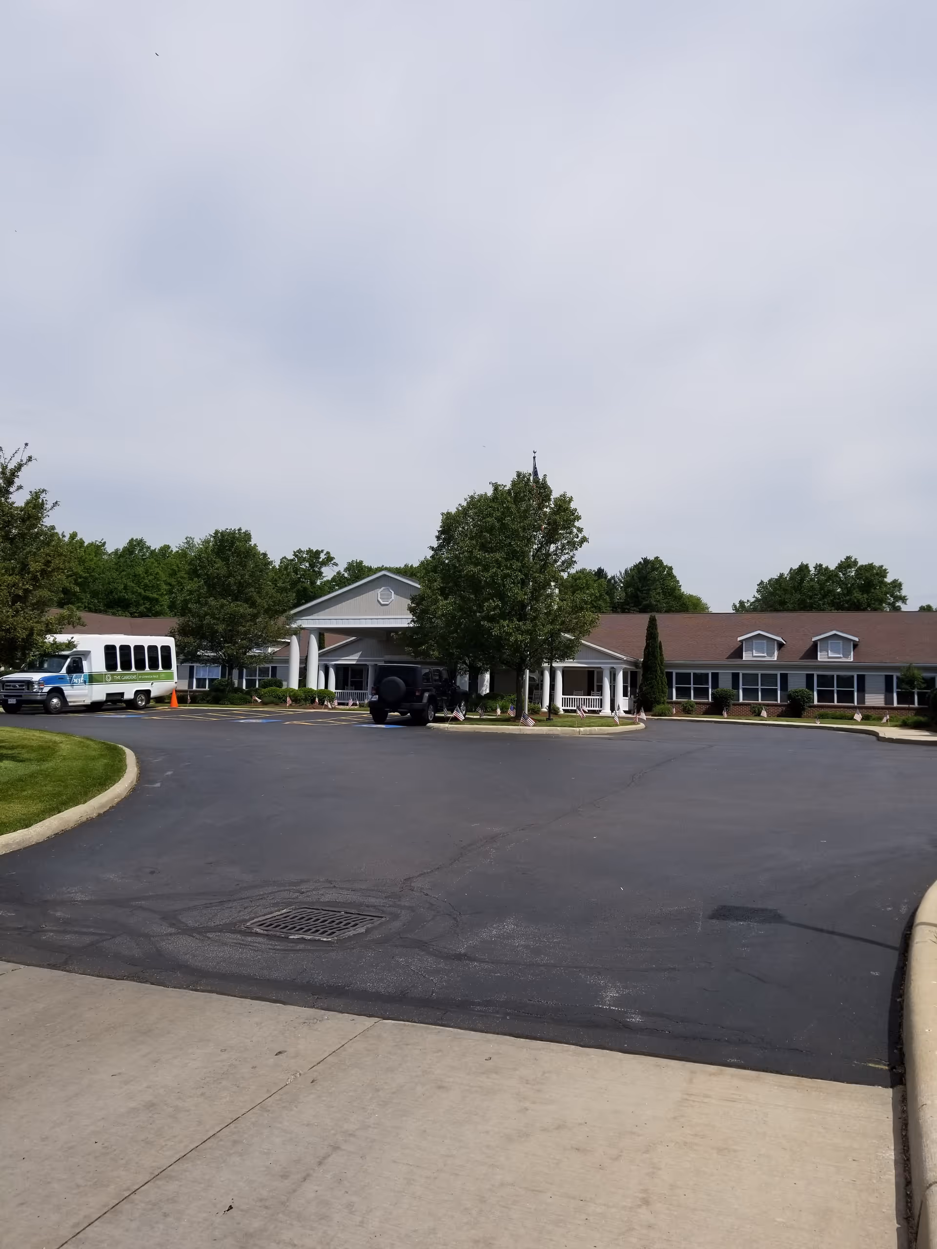 Exterior view of The Gardens at Cuyahoga Falls senior living facility showing a large parking area in front, a single-story building with a peaked roof, white columns at the entrance, surrounded by trees and greenery under a cloudy sky.