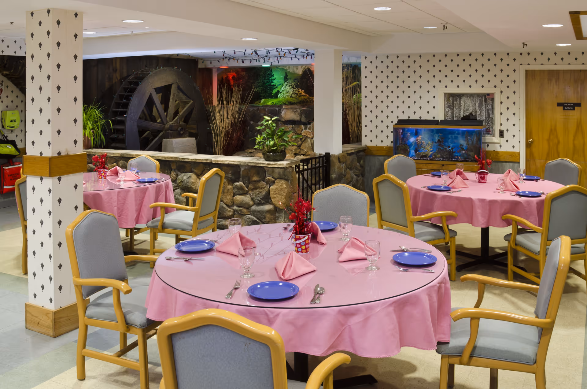 Dining room with round tables covered in pink tablecloths set with blue plates and napkins, surrounded by chairs, with an aquarium and a decorative water wheel in the background.