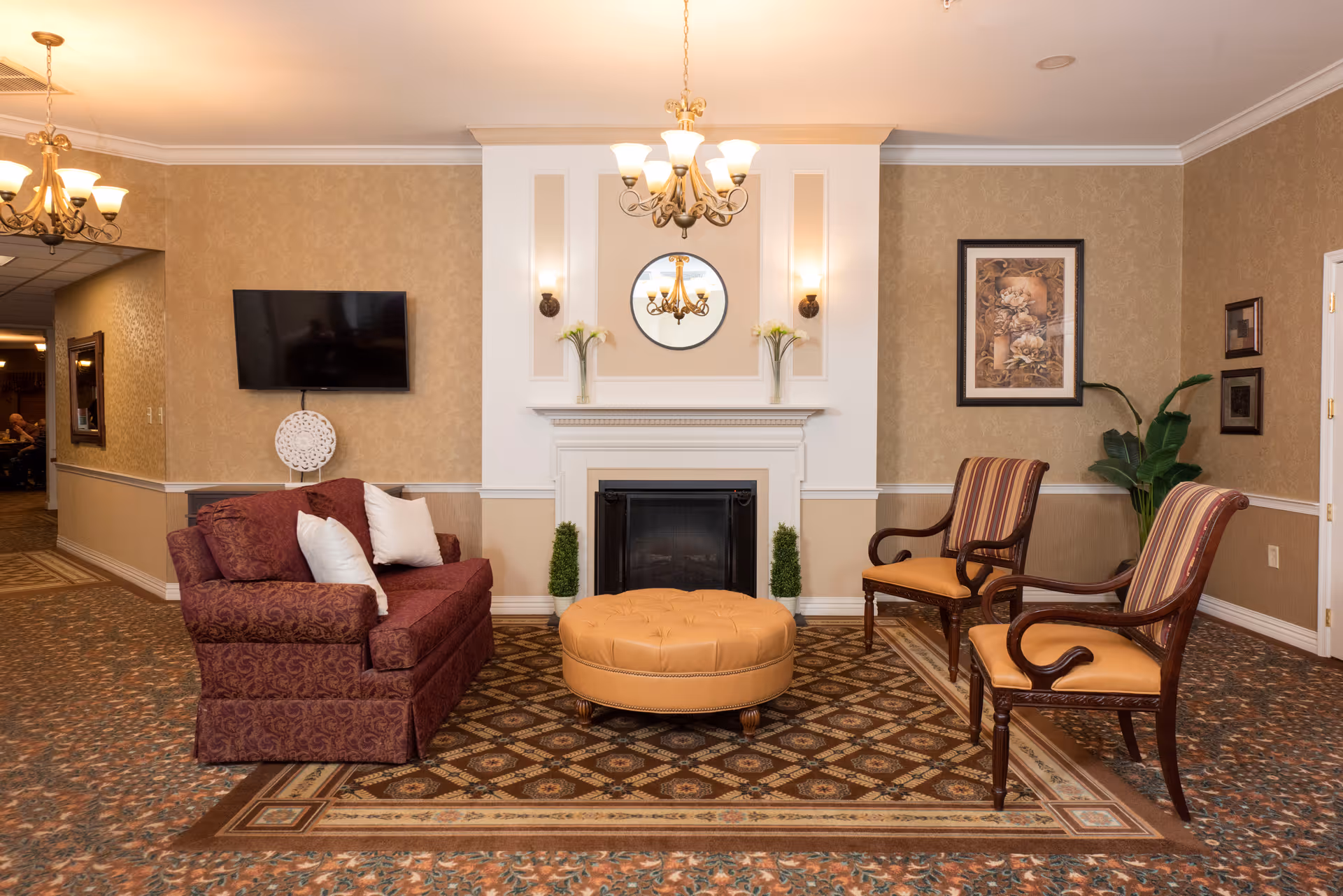 A cozy senior living facility common area featuring a maroon patterned sofa with white pillows, two wooden armchairs with striped cushions, and a round tan ottoman in the center. The room has a fireplace with decorative plants and a round mirror above it. There is a wall-mounted flat-screen TV on the left side and framed artwork on the right wall. The space is warmly lit with chandeliers and wall sconces, and the floor is covered with patterned carpet.