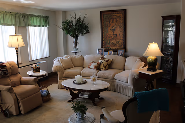 A cozy living room with a beige sofa adorned with decorative pillows, a beige recliner chair, a wooden coffee table with candles and a glass bowl, and two side tables with lamps. There is a large framed artwork on the wall behind the sofa, a tall plant in a vase, and a glass display cabinet in the corner. The room has a window with green valance curtains letting in natural light.