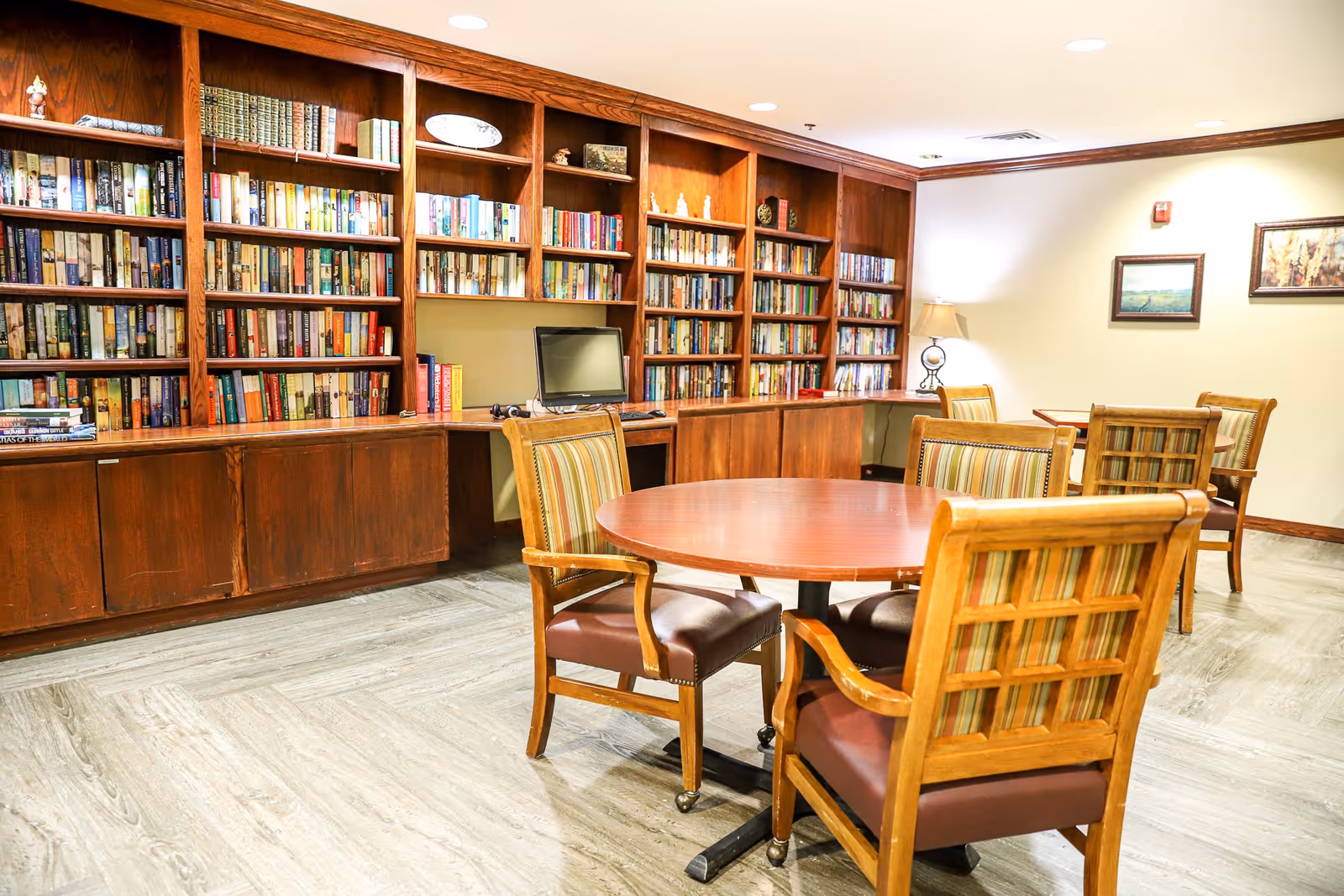 A cozy library room with wooden bookshelves filled with books along the back wall. In front, there are round wooden tables surrounded by wooden chairs with striped cushions. A computer monitor is placed on the desk integrated into the bookshelves. The room is warmly lit with a table lamp and framed artwork on the walls.