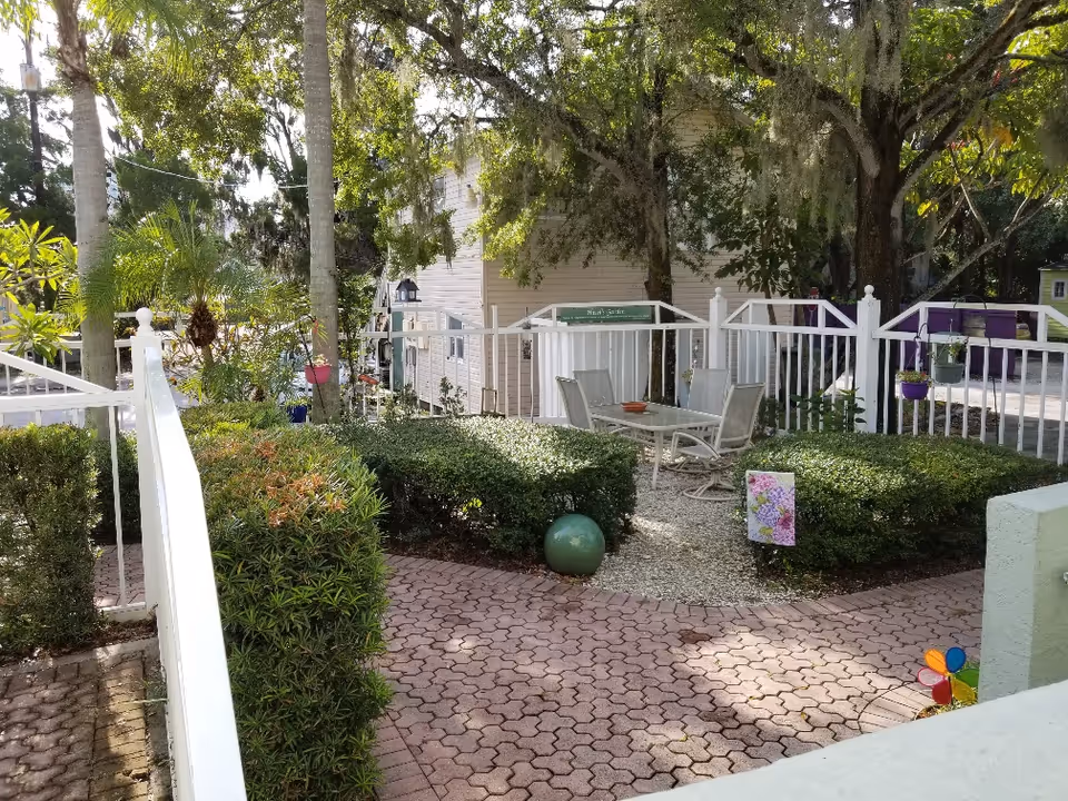 Outdoor garden area with a white fence surrounding a small seating area that has a glass table and four chairs. The garden features trimmed bushes, trees, and various plants. A paved walkway leads to the seating area, and there is a colorful pinwheel decoration on the right side.