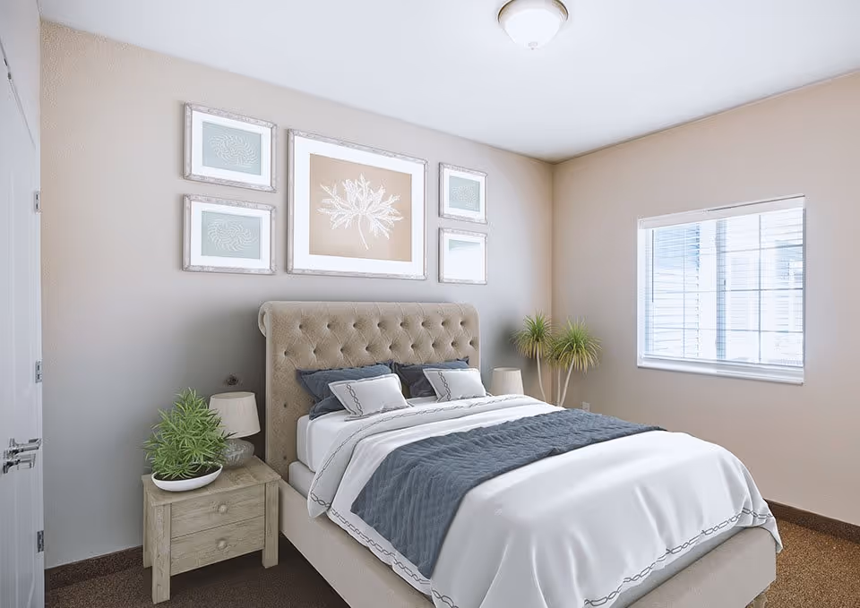 A neatly made bed with a tufted beige headboard in a bedroom. The bed has white sheets, a gray quilted blanket, and several pillows. On the left side of the bed is a wooden nightstand with a lamp and a potted plant. The beige walls have five framed artworks arranged above the headboard. A window with white blinds is on the right wall, allowing natural light into the room.