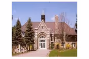 Front exterior view of a large stone building with a steep roof, arched windows, and a central entrance, surrounded by trees and a grassy area.