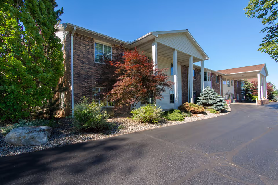 Exterior view of Countryside Manor, a two-story brick and siding building with white columns at the entrance. The driveway is paved and there are various trees and shrubs planted along the building under a clear blue sky.