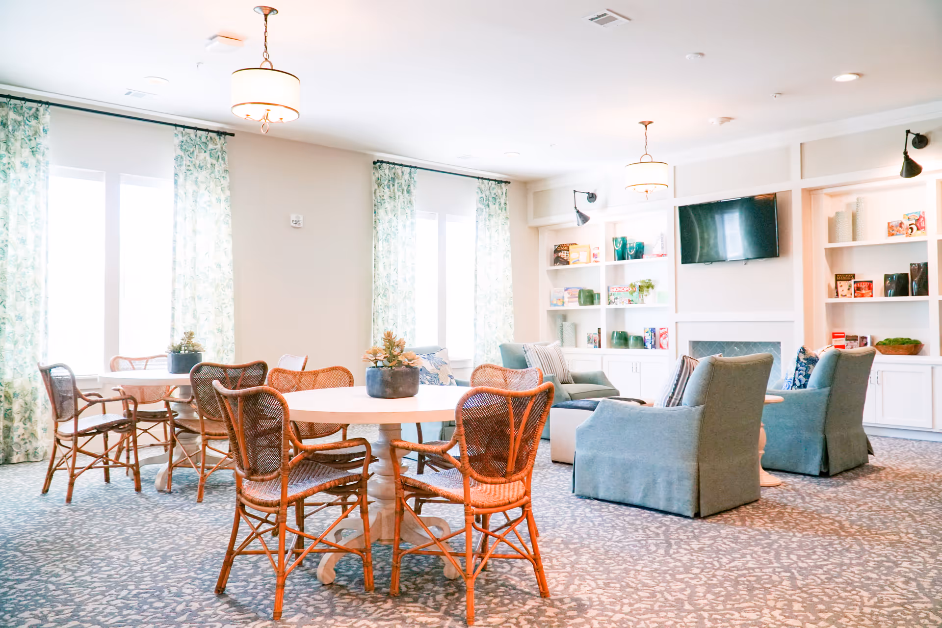 A bright and airy common area with round wooden tables surrounded by wicker chairs, light blue upholstered armchairs, patterned carpet, large windows with floral curtains, built-in white shelves holding board games and decorative items, and a wall-mounted flat screen TV above a fireplace.