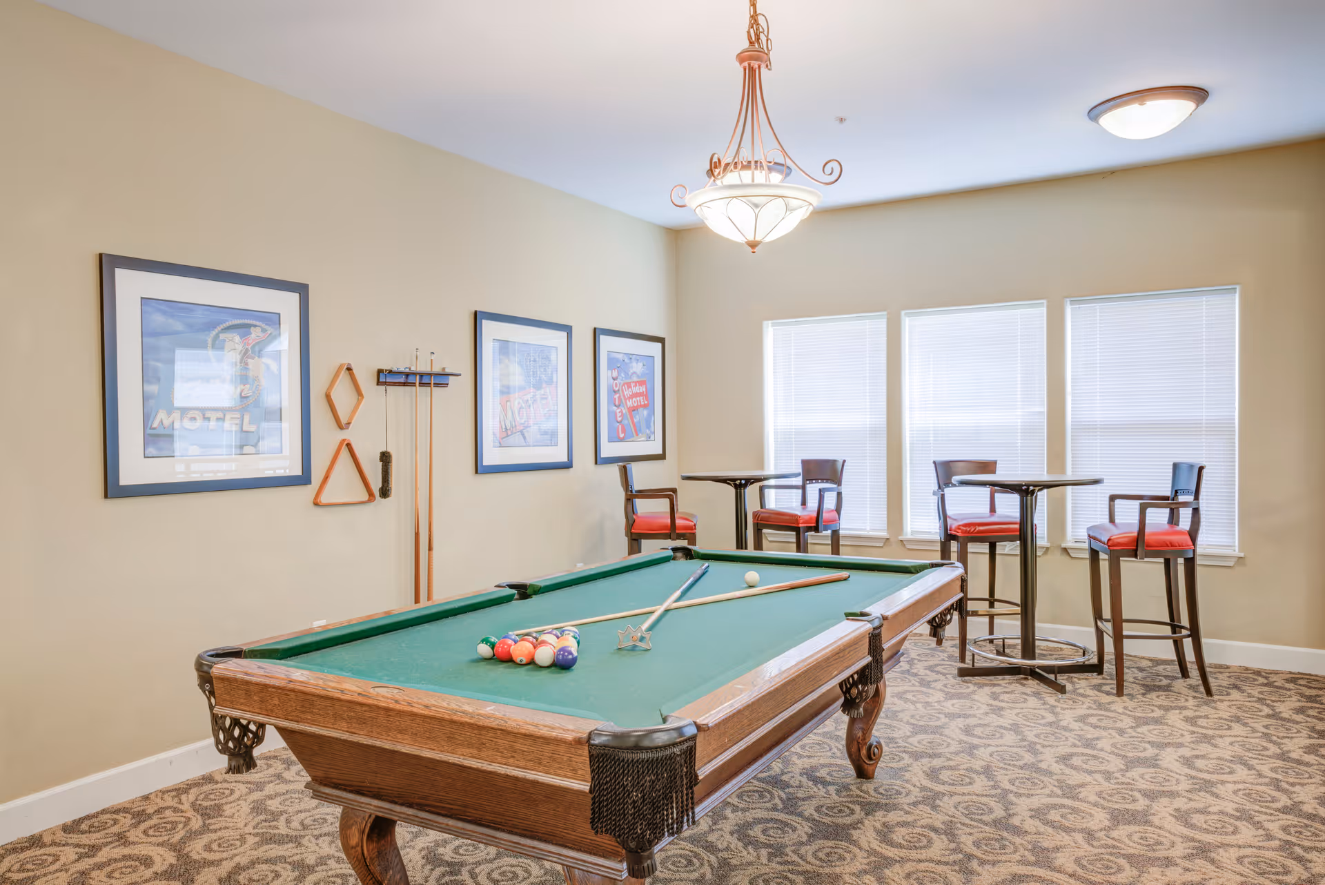 A well-lit game room featuring a green felt pool table with pool balls and cues arranged on it. The room has beige walls adorned with three framed vintage motel-themed pictures and a rack holding pool cues and triangle racks. There are three tall tables with red cushioned chairs near three windows with closed blinds. The floor is carpeted with a patterned design, and two ceiling lights illuminate the space.