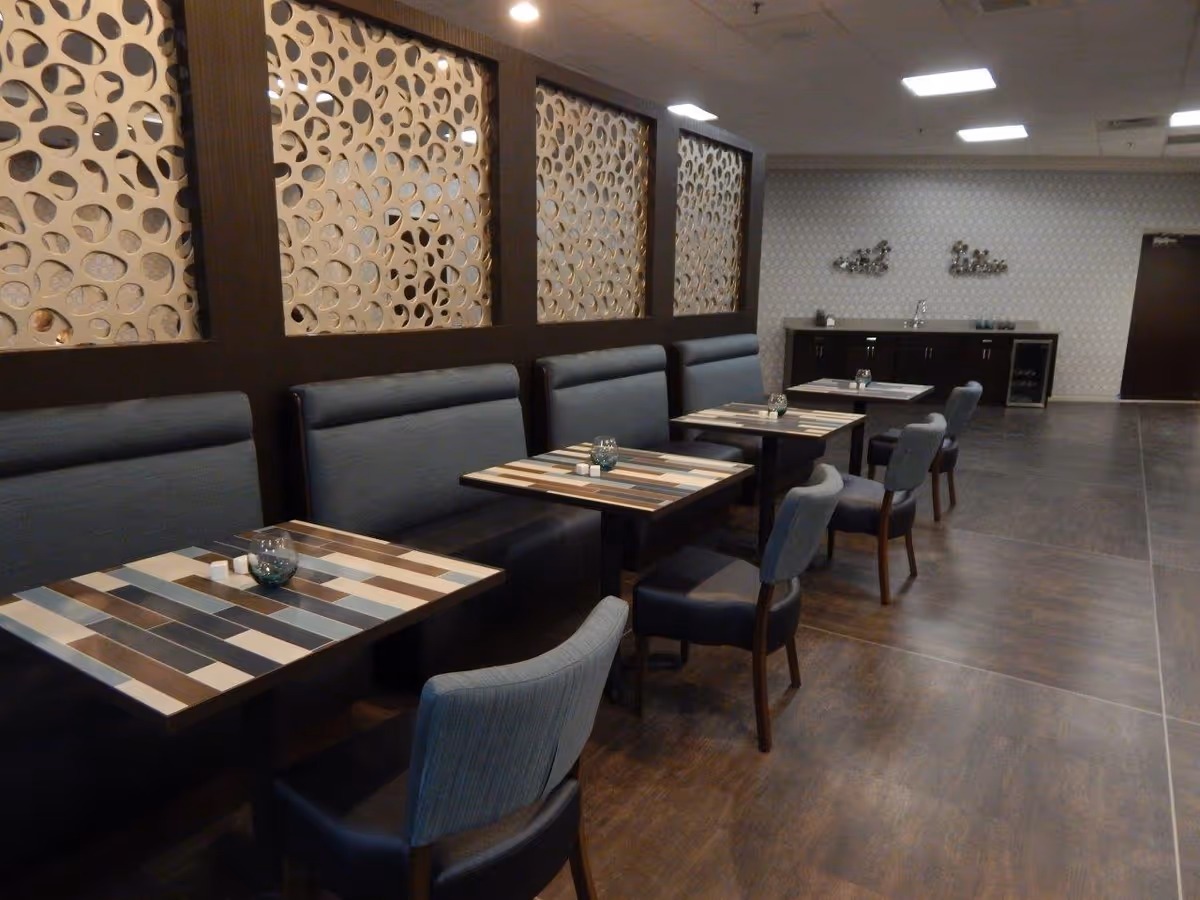 Interior view of a dining area with several tables and chairs arranged in a row. The tables have a striped pattern with various shades of brown, beige, and blue. There are cushioned bench seats along the wall with decorative panels featuring circular cut-out designs. The floor is a dark wood finish, and the back wall has a small counter with cabinets and decorative wall art.