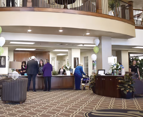 Lobby of a senior living facility showing a reception desk, several people standing and a few balloons.