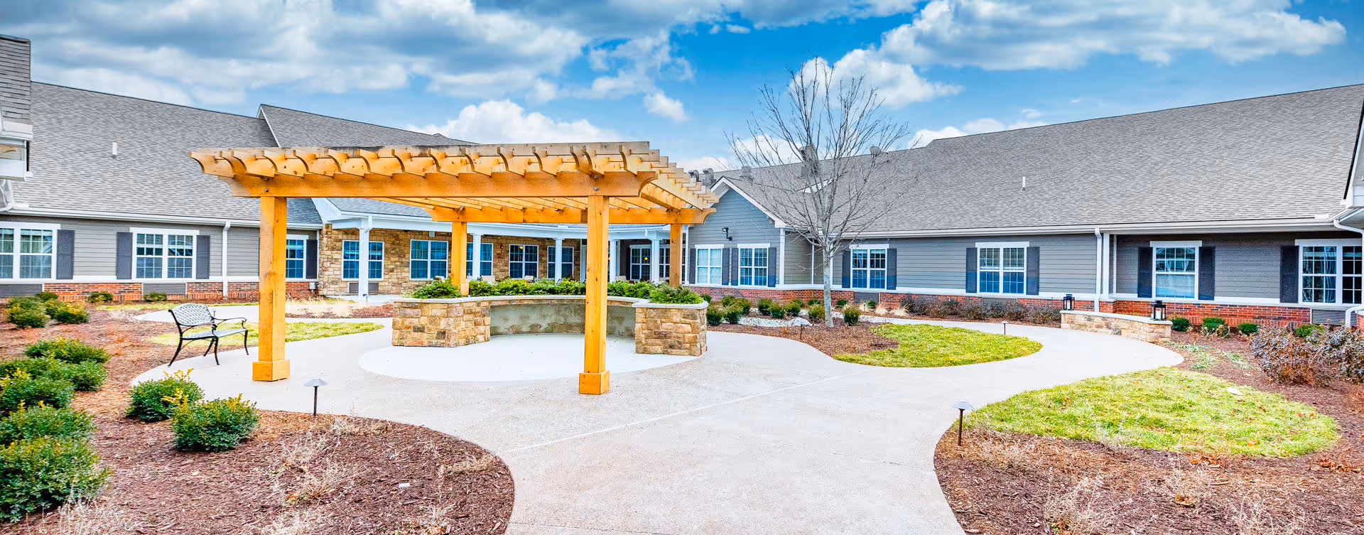 Outdoor courtyard area at Bickford of Canton featuring a wooden pergola with stone seating underneath, surrounded by landscaped garden beds, a curved concrete walkway, a bench, and a large single-story building with multiple windows in the background under a partly cloudy sky.