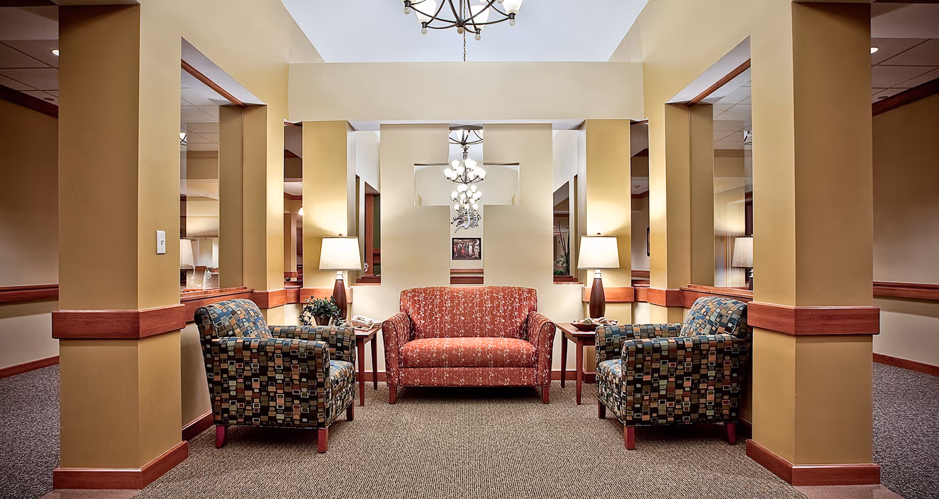 A cozy seating area in a senior living facility with a red patterned loveseat centered between two multicolored patterned armchairs. Each side of the loveseat has a wooden side table with a lamp and decorative items. The walls are painted beige with wooden trim, and there are multiple mirrors reflecting the chandelier lights above.