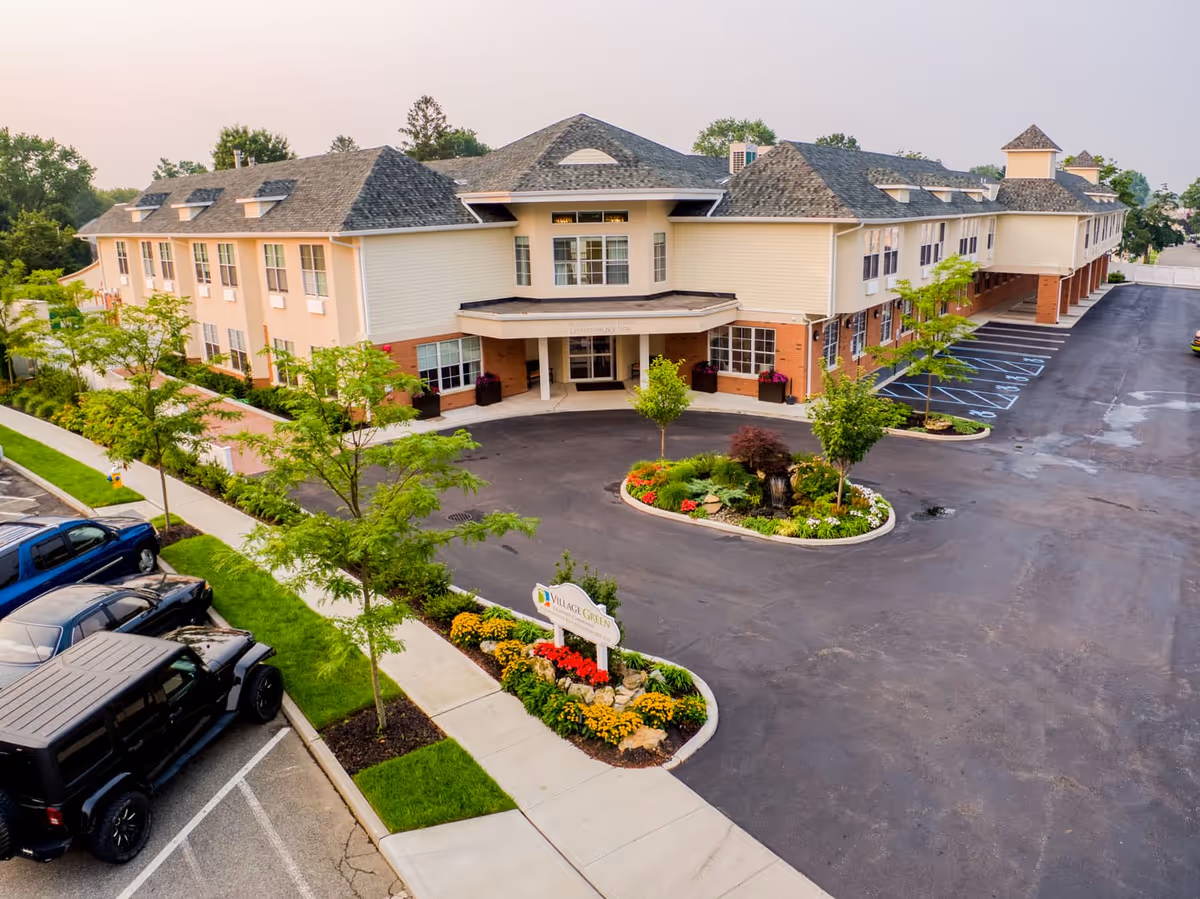 Aerial view of a large two-story senior living facility named Village Green, featuring a landscaped entrance with flowers and trees, a driveway, and parking spaces with several vehicles parked.