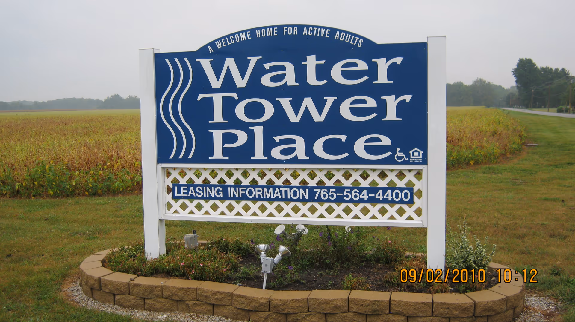 Outdoor sign for Water Tower Place, a community described as 'A Welcome Home for Active Adults,' with leasing information phone number 765-564-4400, set in a landscaped area with grass and plants, and a field and road in the background.