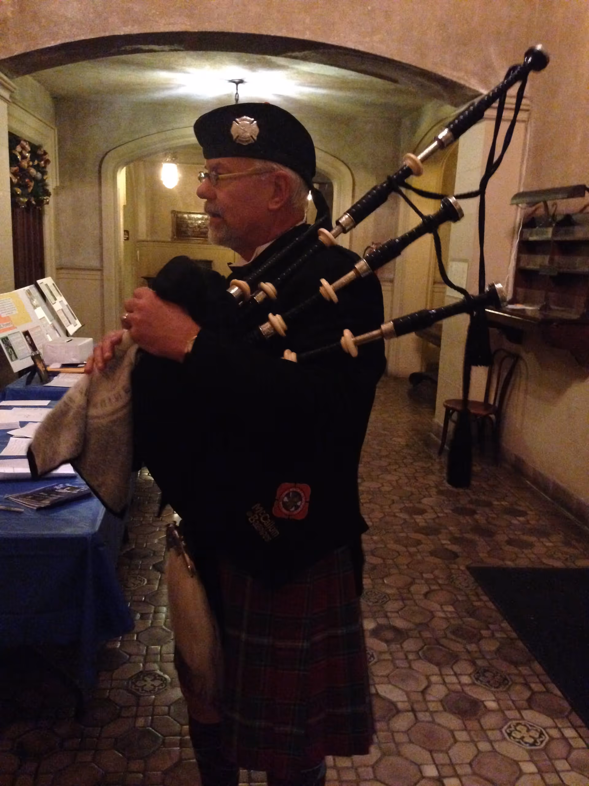An elderly man wearing traditional Scottish attire, including a kilt and a black hat with an emblem, playing the bagpipes indoors in a hallway with tiled flooring and arched ceilings.