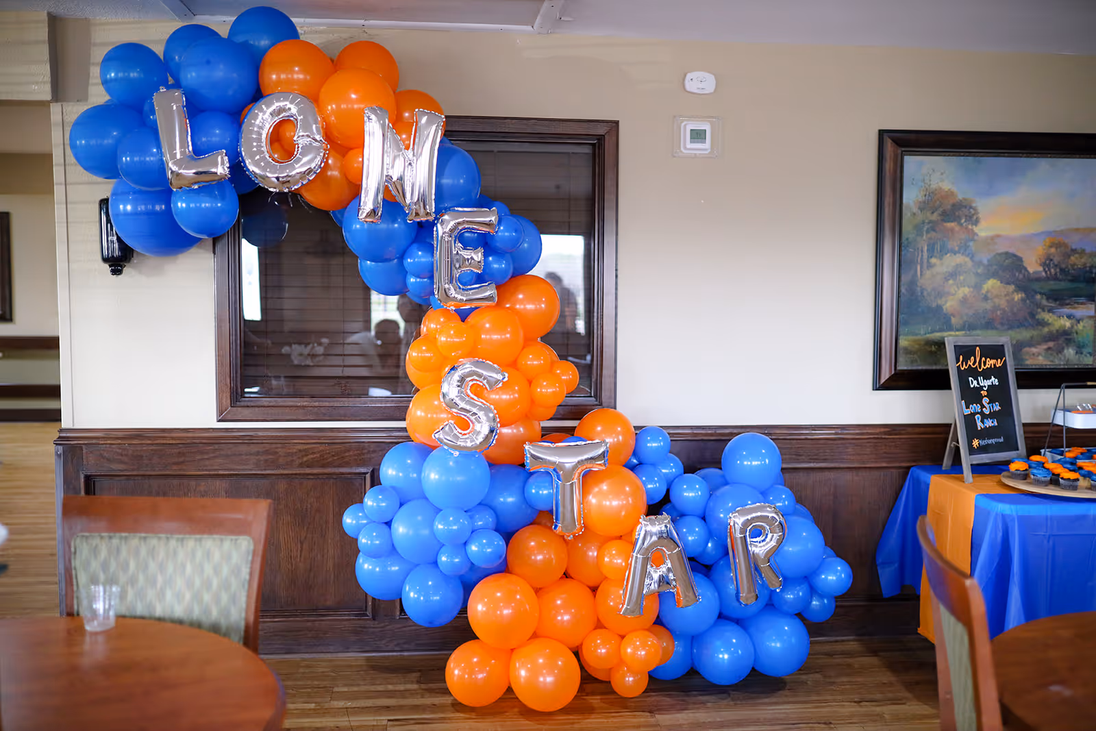 A colorful balloon arrangement in blue and orange spelling out 'LONE STAR' in silver letter balloons is displayed indoors against a wall with wood paneling. To the right, there is a table covered with a blue and orange tablecloth, holding a small chalkboard sign that says 'Welcome Dr. Ulyette Lone Star Ranch' along with some cupcakes. The setting appears to be a common area or dining space in a facility.