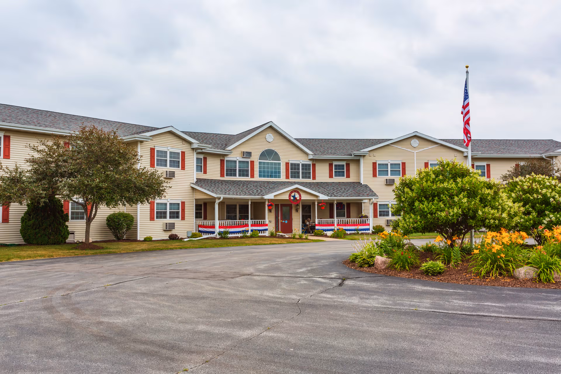 Exterior view of The Cottages at Meadowlands Senior Living building with beige siding, red shutters, a covered entrance decorated with a wreath, an American flag on a flagpole, and landscaped greenery including trees and flowering plants.