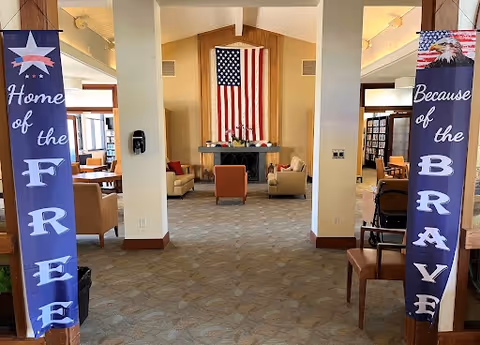Interior view of a common area in a senior living facility with an American flag hanging on the back wall above a fireplace. The room has beige armchairs arranged around the fireplace and bookshelves along the walls. Two vertical banners hang on wooden pillars at the entrance, one reading 'Home of the FREE' and the other 'Because of the BRAVE' with patriotic imagery.