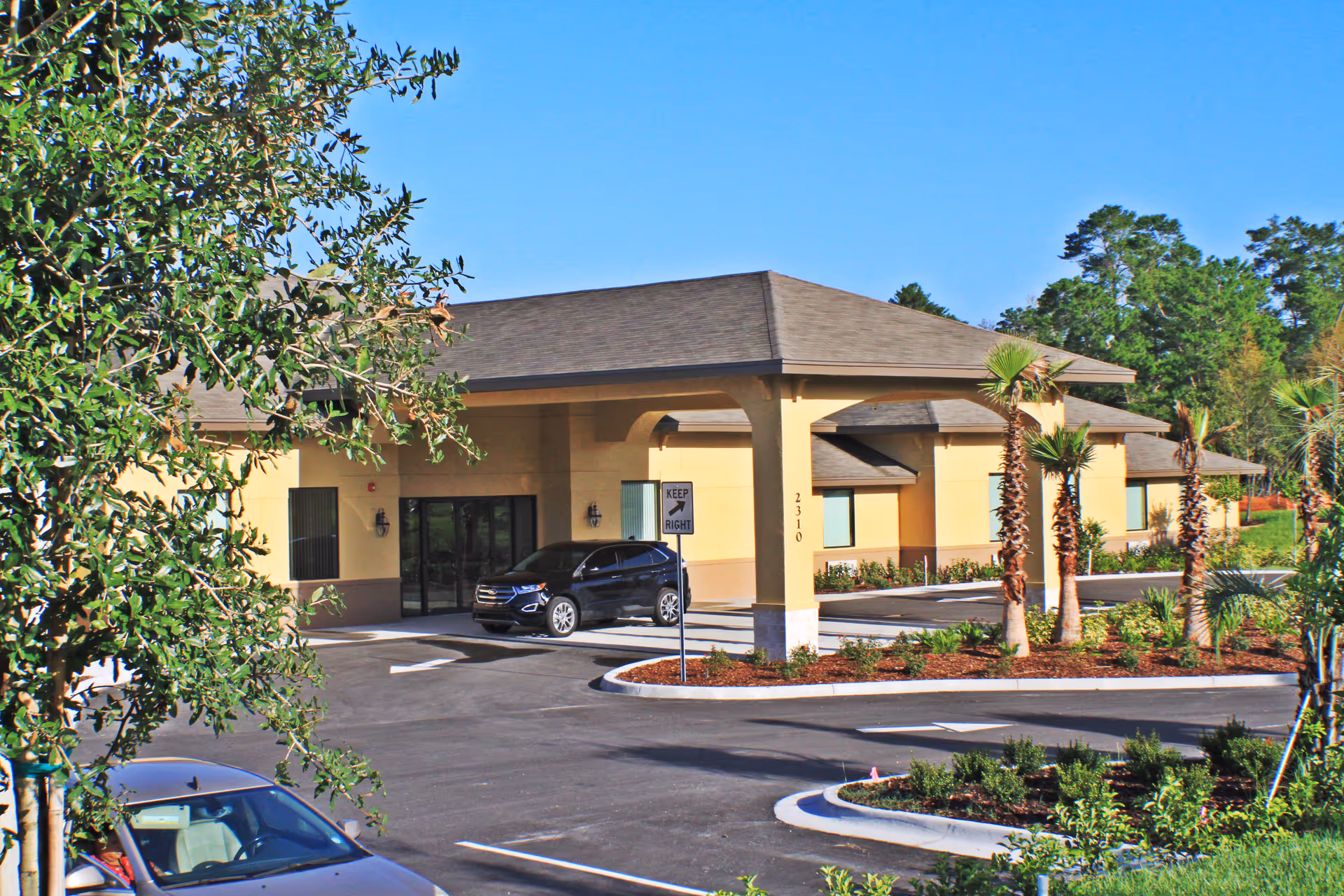 Exterior view of a single-story assisted living facility building with a covered entrance, surrounded by landscaped greenery and palm trees, with a few parked cars in the driveway under a clear blue sky.
