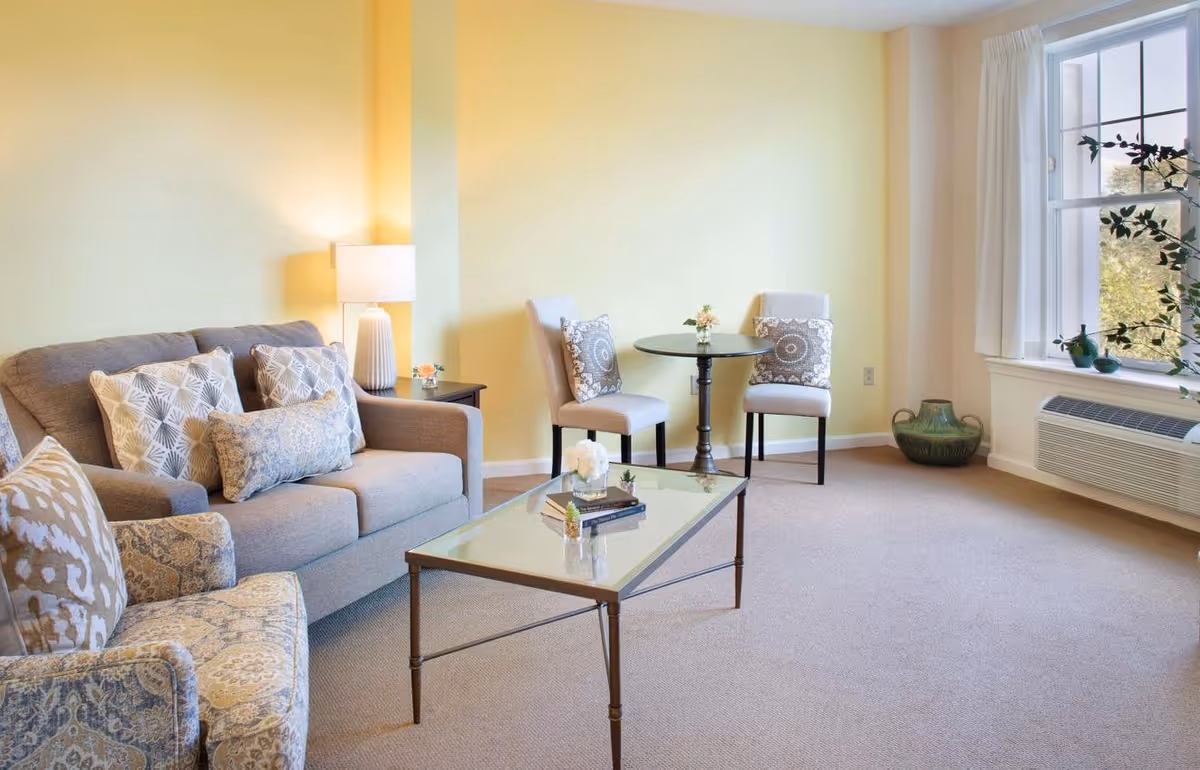 A cozy living room with a gray sofa adorned with patterned cushions, a matching armchair, a glass coffee table with decorative items, two beige chairs with patterned pillows around a small round table, a side table with a lamp, and a large window with white curtains letting in natural light.